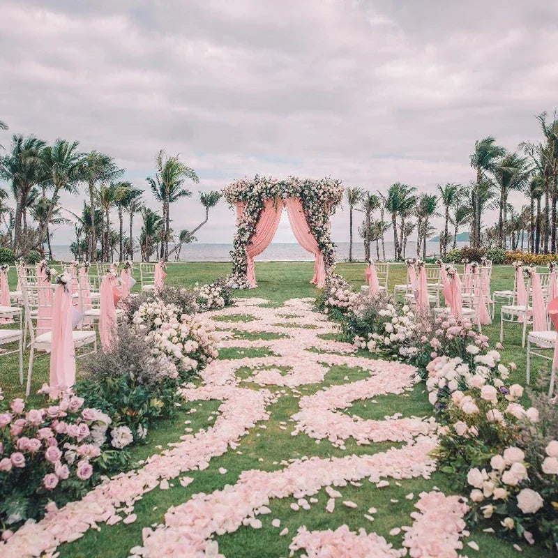 A green aisle decorated with pink rose petals leading to an archway surrounded by greenery and palm trees for a wedding.