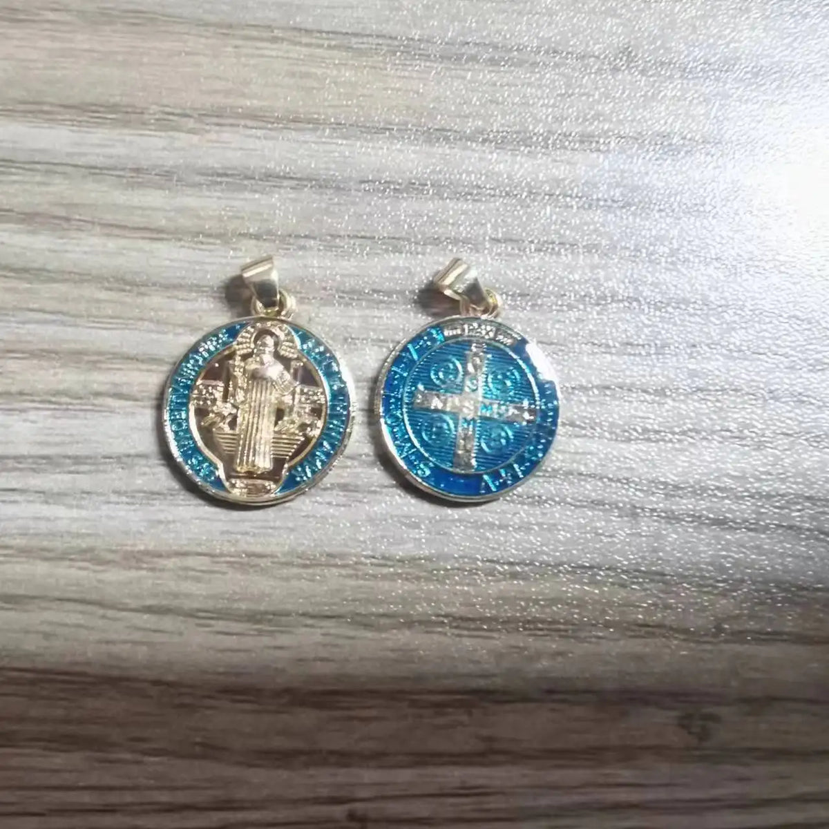A pair of silver-toned religious pendants featuring St. Benedict imagery and a cross design on blue enamel.