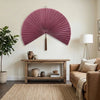 A large, pink woven bamboo fan hangs on a white wall above a wooden shelf in a living room setting.