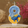 A birthday candle shaped like the number 8 featuring a blue dinosaur design and glittery blue and orange icing.
