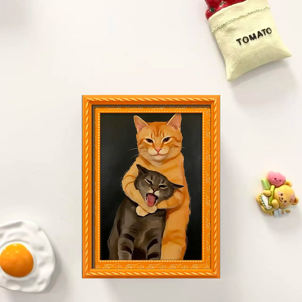 A framed painting featuring an orange cat hugging a black cat, displayed on a white surface.