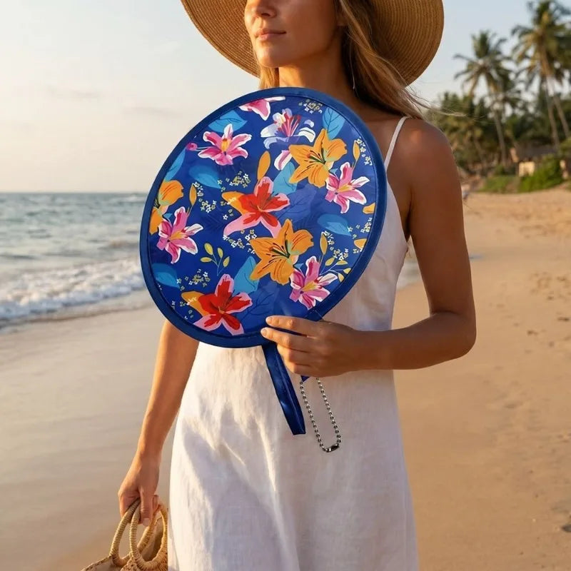 A woman holds a blue floral pocket fan with orange and pink flowers on a beach.