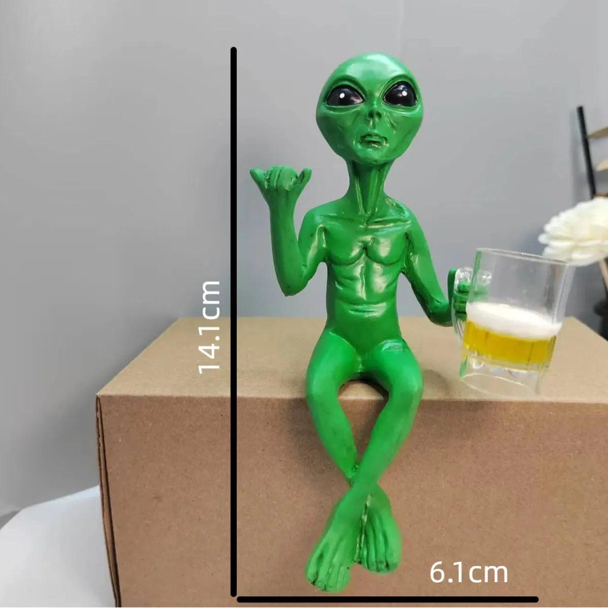 A green resin alien statue with an arm holding a beer mug, displayed on a cardboard box.