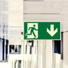 A green exit safety sign with a silhouette of a person walking and a downward arrow on a light-colored wall.