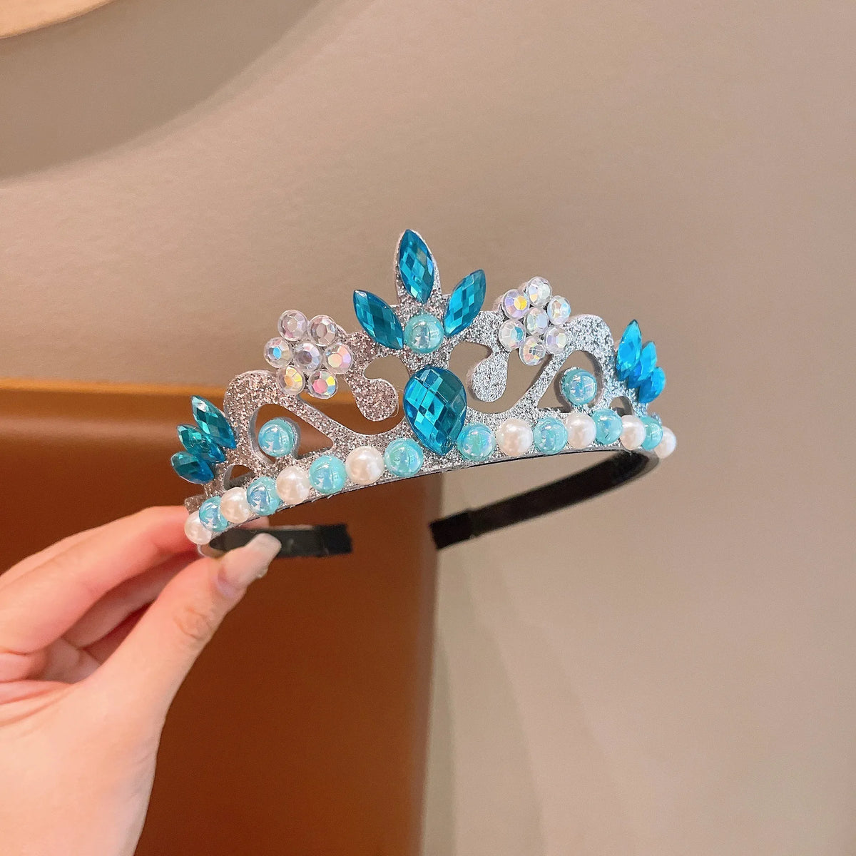 A silver crown headband adorned with blue and clear faux gems and pearls being held by a hand.