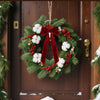 A festive green pine wreath decorated with red berries and white velvet Christmas bows hanging on a wooden door.