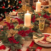A festive holiday arrangement featuring green pine branches, red holly berries, and glowing candles on a wooden table.