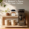 A handwoven knitted coffee cup cozy with a smiling doll decoration sits next to a coffee grinder and a mug on a wooden shelf.