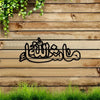 Black Arabic calligraphy design on a wooden wall with green foliage and grass in the foreground.