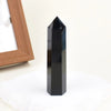 A black natural obsidian crystal obelisk point standing on a white surface.