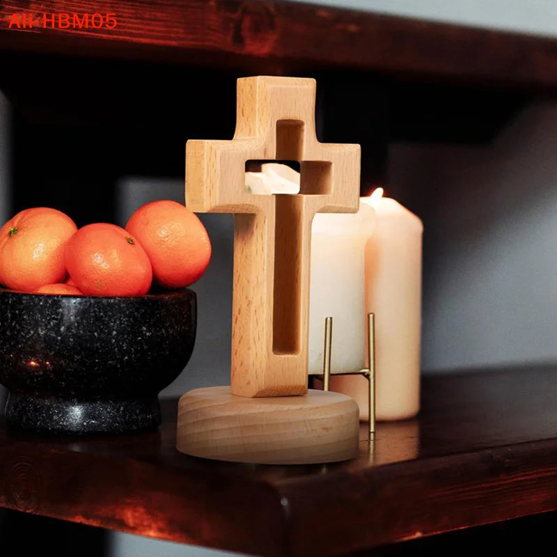A handcrafted wooden cross standing ornament placed on a dark bowl next to a lit candle on a wooden shelf.