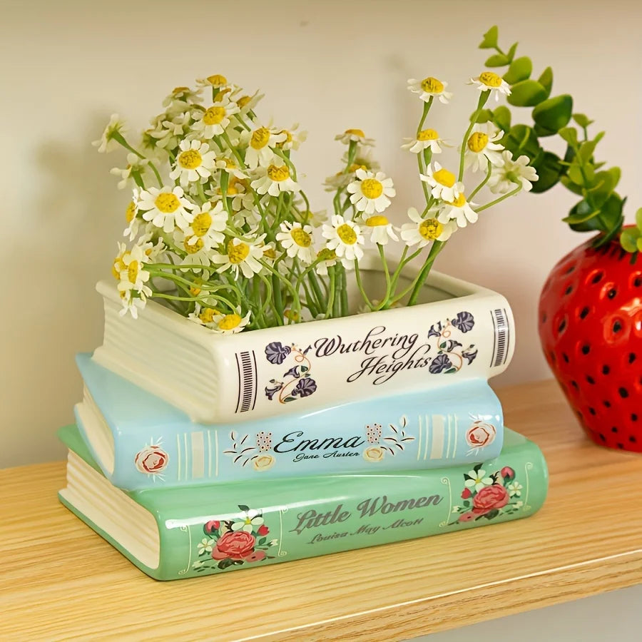 Une pile de trois livres en céramique vert clair et blanc avec des motifs floraux, surmontée d'un vase de marguerites blanche