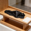 A black, draped reclining female statue sculpture is displayed in a wooden box on a white surface.