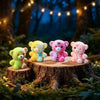 A set of four colorful resin bear figurines in yellow, blue, pink, and white are displayed on a wooden stump in a forest sett