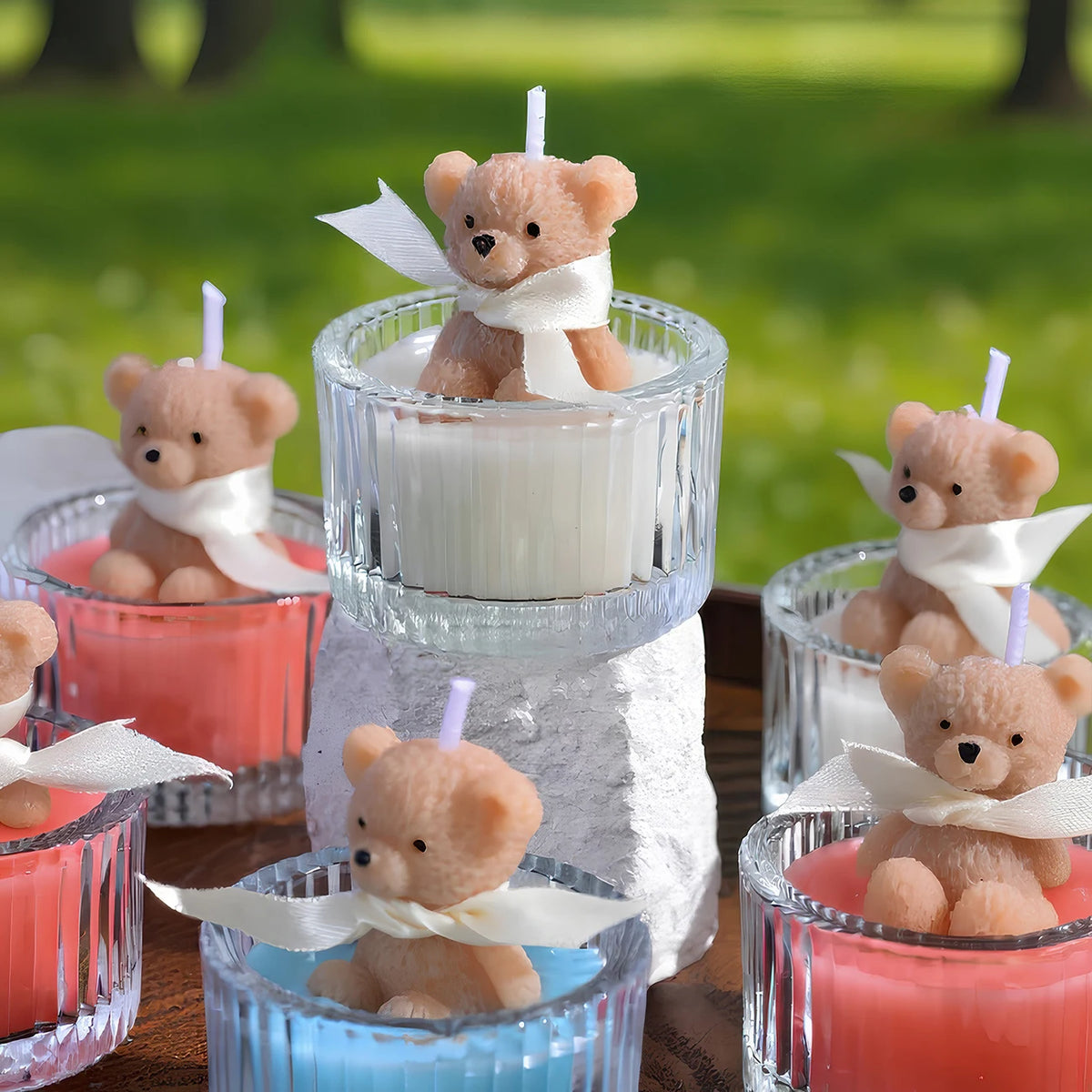A collection of soy wax teddy bear candles in glass jars with ribbons, featuring pink, orange, and blue colors.