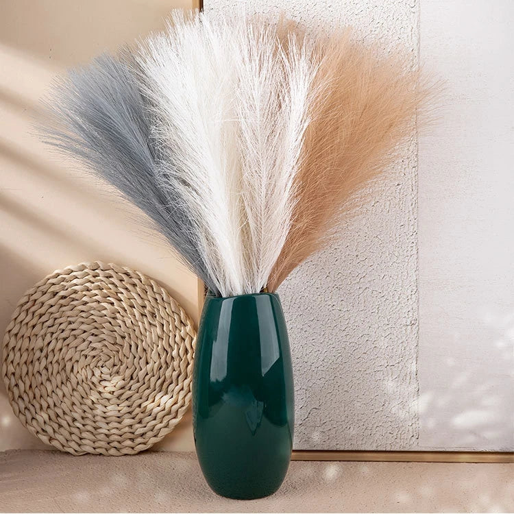A vase holds artificial pampas grass stems in shades of gray, white, and tan against a light background.