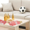 A table setting with glass bottles, a soccer ball, and red and white paper popcorn bags on a light wooden table.