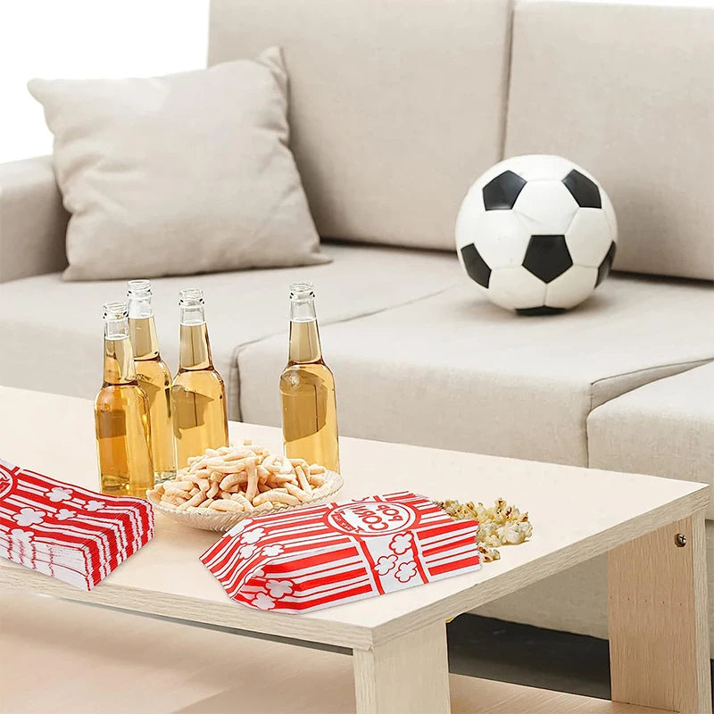 A table setting with glass bottles, a soccer ball, and red and white paper popcorn bags on a light wooden table.
