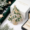 A gold-toned rhinestone Christmas tree brooch pin displayed on a white box with green velvet and pine branches.