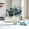 A white ceramic vase filled with silvery-blue foliage and small white flowers on a marble countertop.