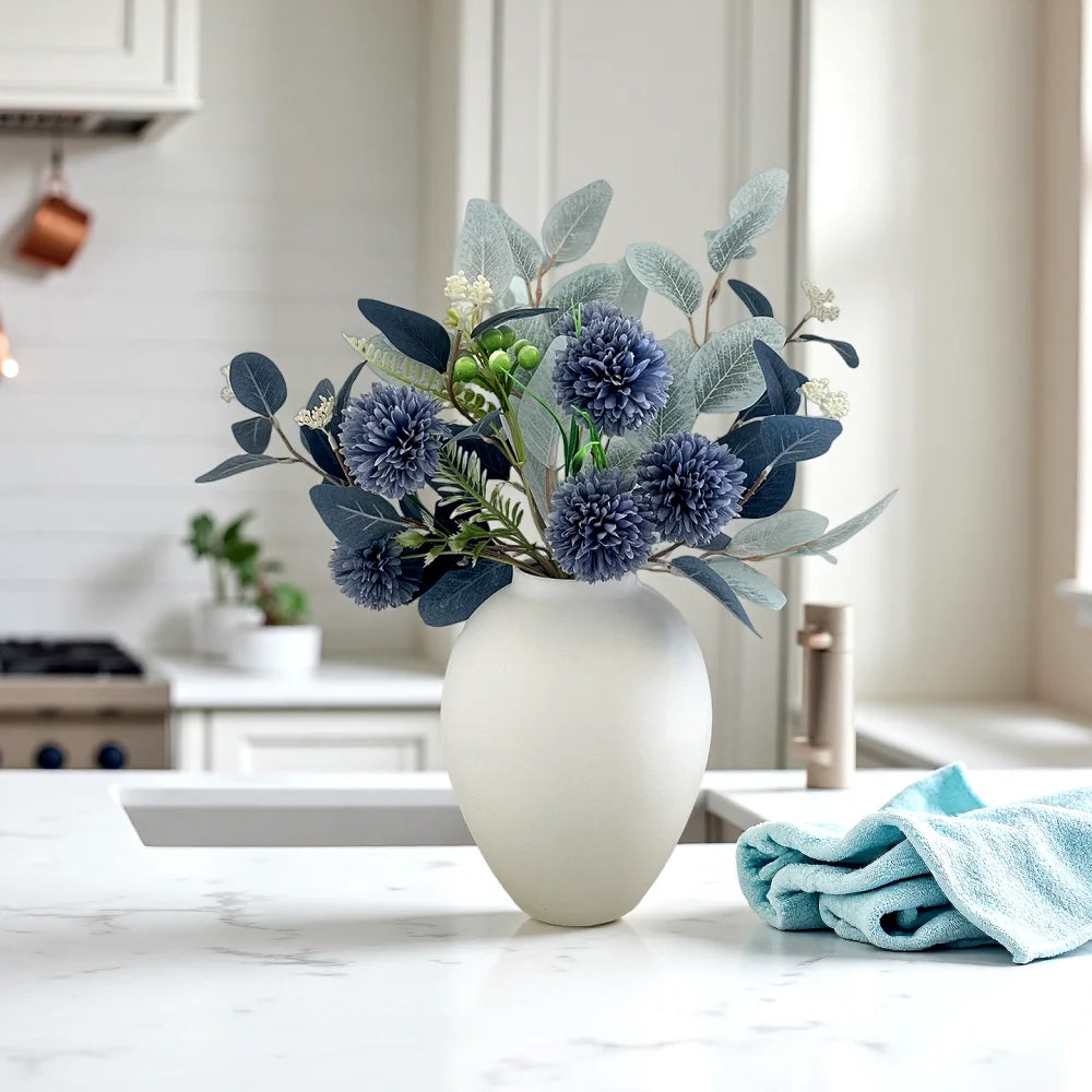 A white ceramic vase filled with silvery-blue foliage and small white flowers on a marble countertop.