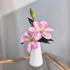 Three pink and white artificial lilies with buds in a white vase on a wooden tray.