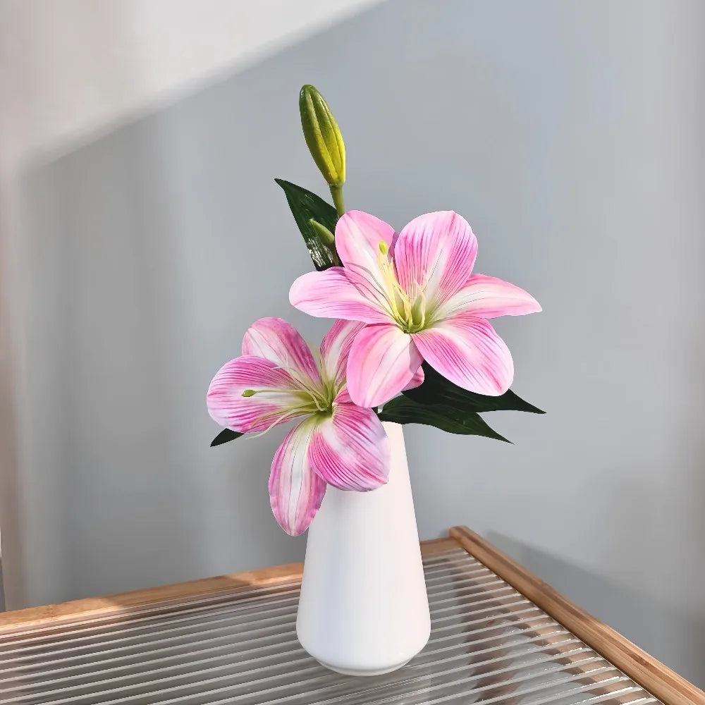 Three pink and white artificial lilies with buds in a white vase on a wooden tray.