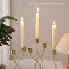 A set of three gold-toned iron candlesticks with white candles displayed on a white surface.