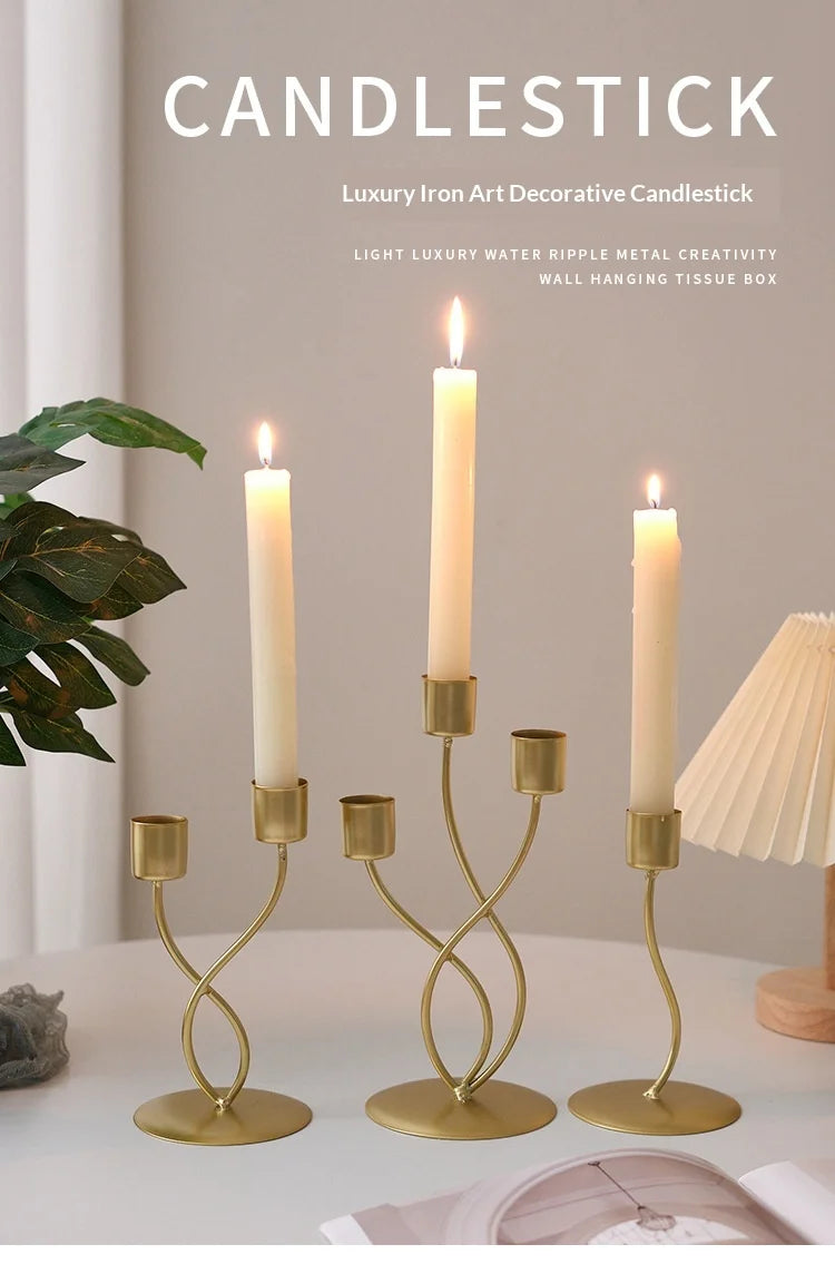 A set of three gold-toned iron candlesticks with white candles displayed on a white surface.