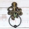 A bronze octopus skull door knocker featuring a cast iron ring, displayed on a white wooden surface.