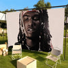 A portrait of a man with dreadlocks and a bandana displayed on a large white wall tapestry outdoors.