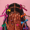 A close-up of a woman's hands covering her face, adorned with gold jewelry and colorful braids, against a pink background.