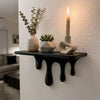 A black drip floating shelf holds a white candle and a potted succulent on a white wall.