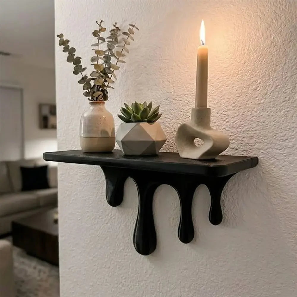 A black drip floating shelf holds a white candle and a potted succulent on a white wall.