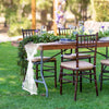 A rustic outdoor wedding table set with dark wood chairs, greenery, and a white embroidered lace runner on a wooden table.