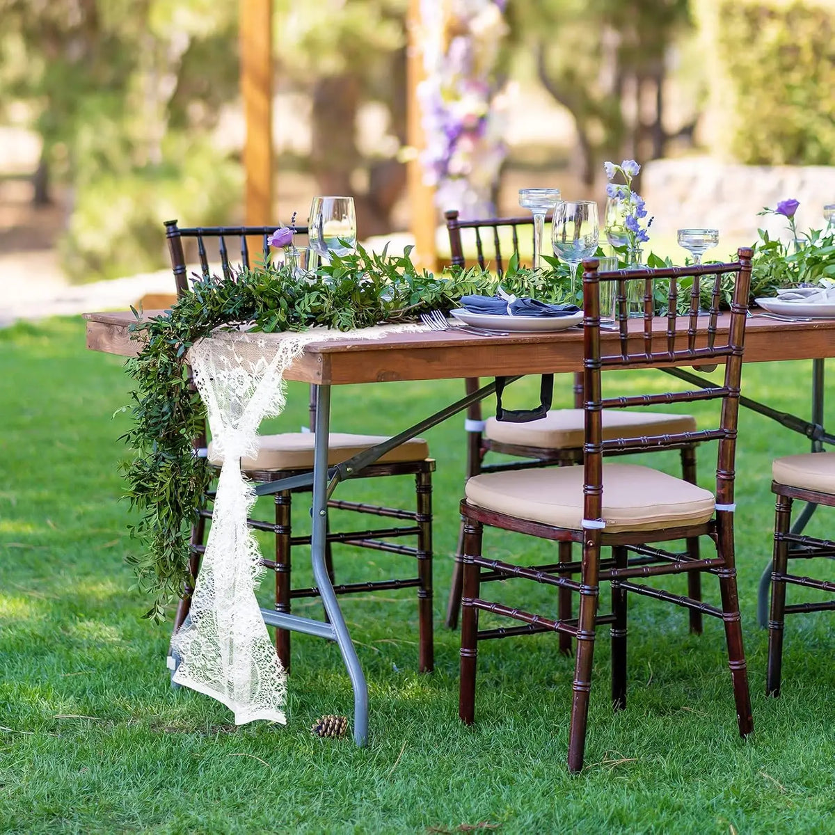 A rustic outdoor wedding table set with dark wood chairs, greenery, and a white embroidered lace runner on a wooden table.