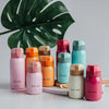A collection of pink, orange, and mint green stainless steel vacuum insulated coffee mugs arranged in front of a large green 
