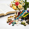 A collection of wooden rosary beads and crucifix necklaces arranged on a white surface with roses and white flowers in the ba