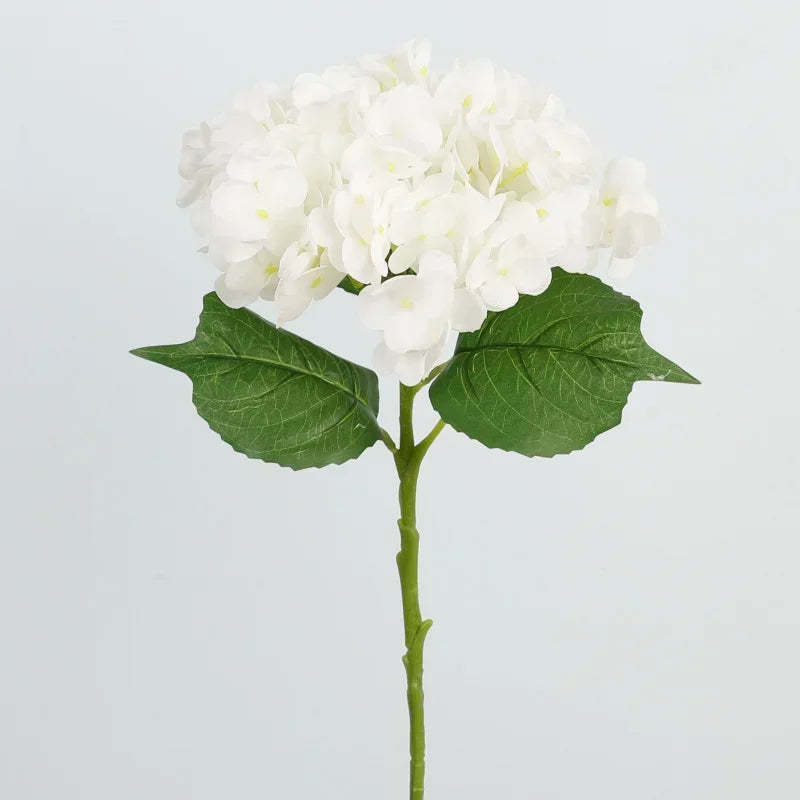 A sprig of white artificial hydrangea flowers on a green stem with large leaves.