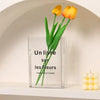 Clear acrylic book vase holding two yellow orange tulips on a white shelf.