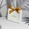 A white paperboard thank you gift bag tied with a gold ribbon, set against a background of palm shadows.
