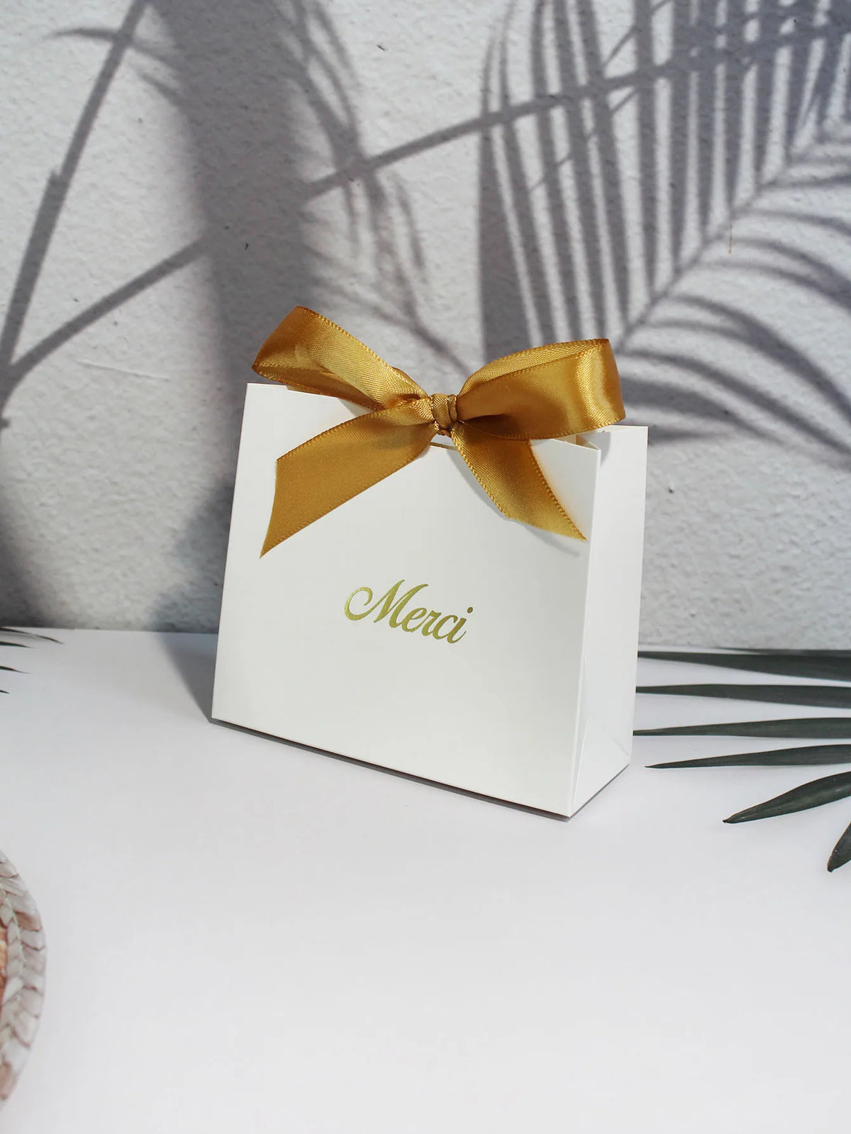 A white paperboard thank you gift bag tied with a gold ribbon, set against a background of palm shadows.