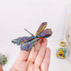 A hand holds a natural crystal tourmaline dragonfly figurine with iridescent purple and green wings.