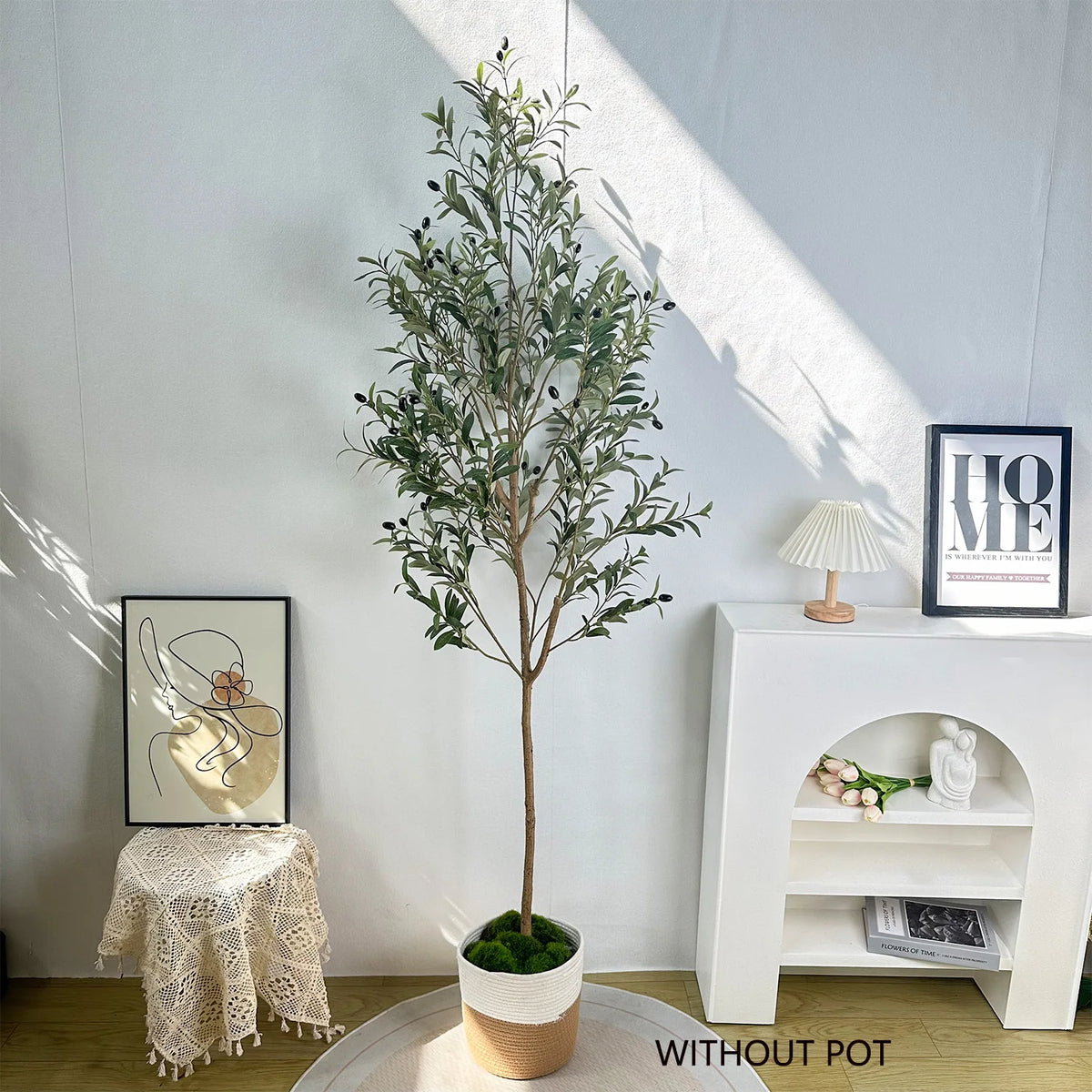 A tall artificial olive tree branches in a pot displayed against a white wall with framed art and a small shelf.