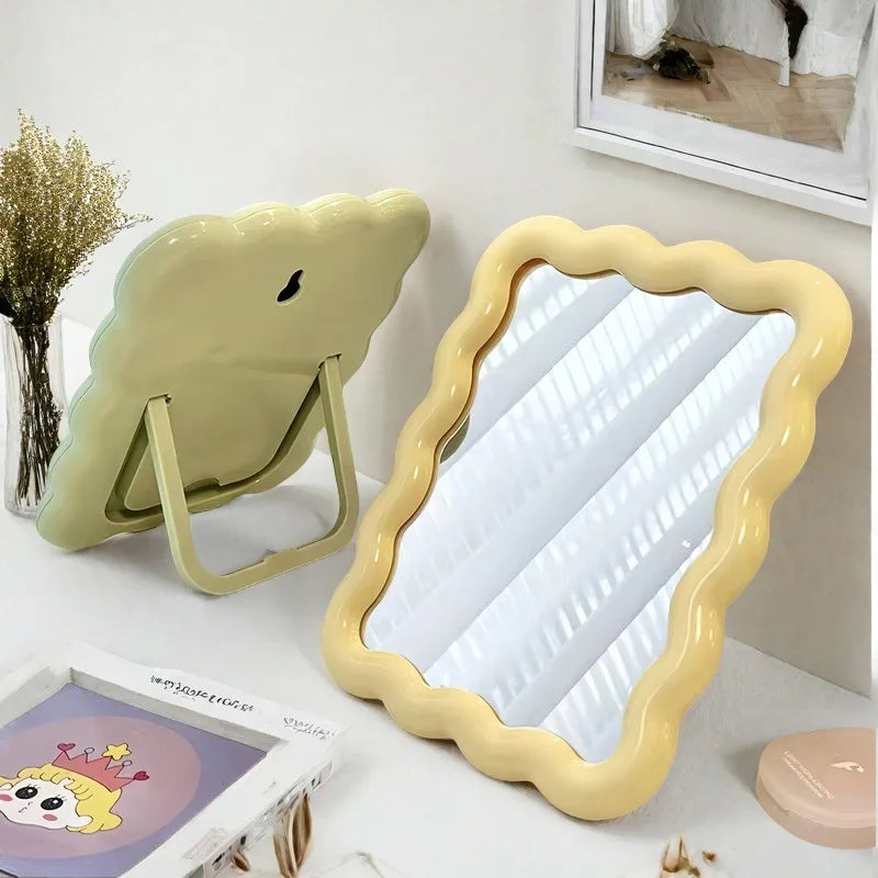 A light green wavy frame makeup mirror with a gold stand is displayed on a white surface.