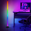 A purple and rainbow RGB floor lamp illuminates a desk setup featuring a laptop and a keyboard.
