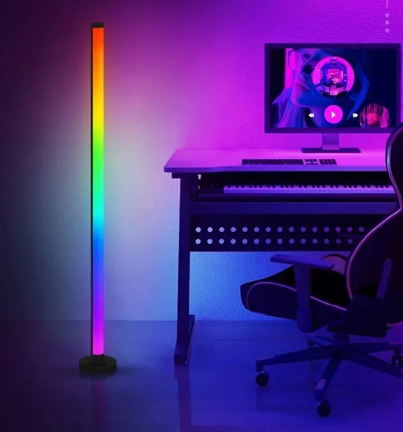 A purple and rainbow RGB floor lamp illuminates a desk setup featuring a laptop and a keyboard.