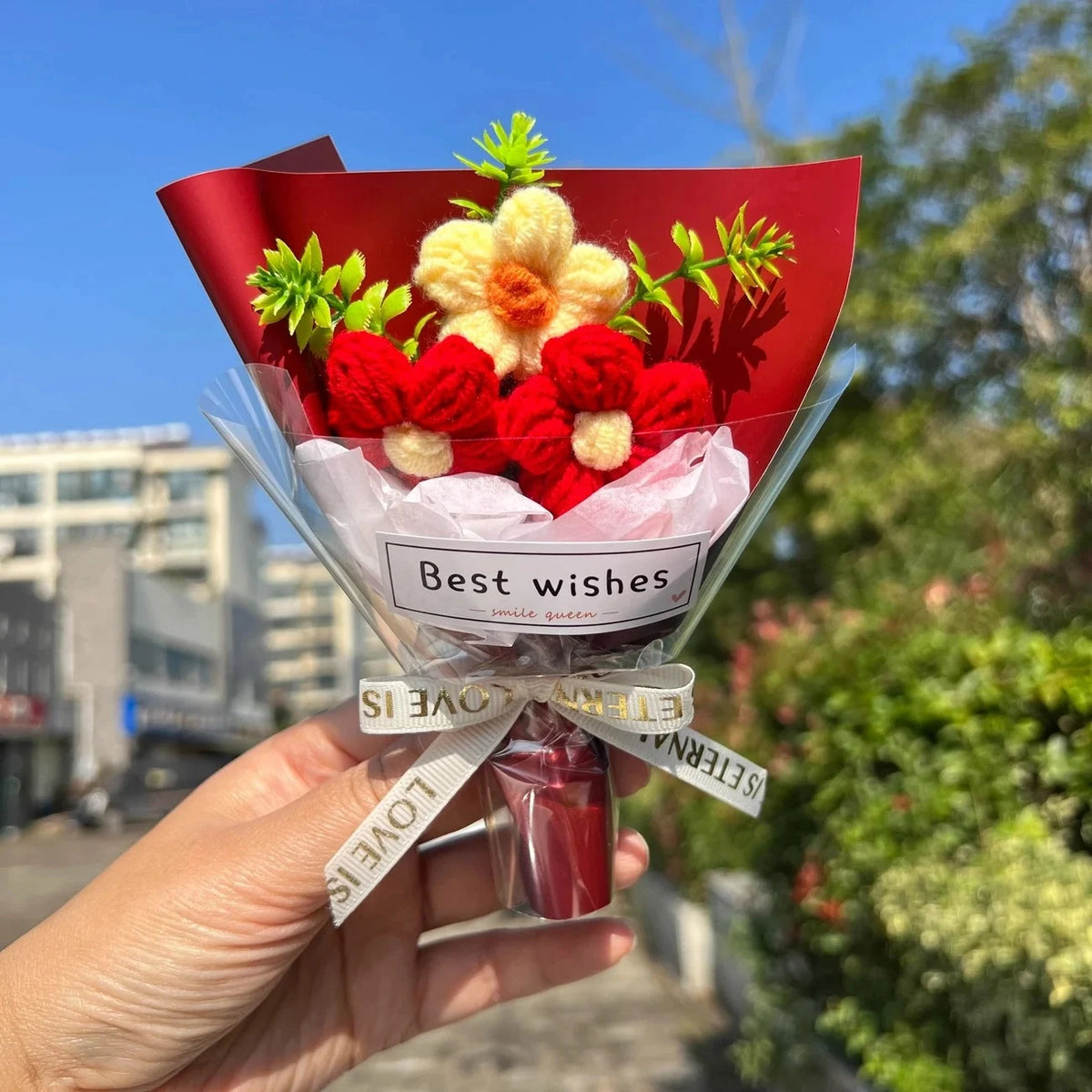 A hand holds a bouquet of red and yellow crochet flowers wrapped in red paper with a ribbon.