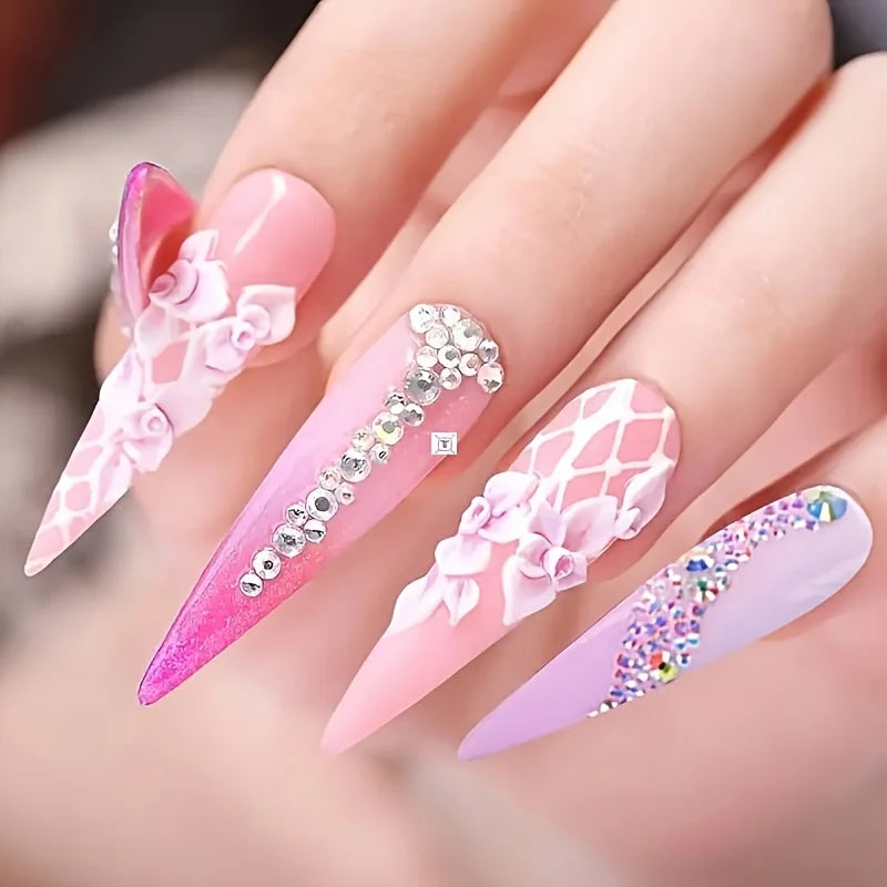 A hand displays various pink and purple acrylic nails decorated with floral designs and glittery micro beads for nail art.
