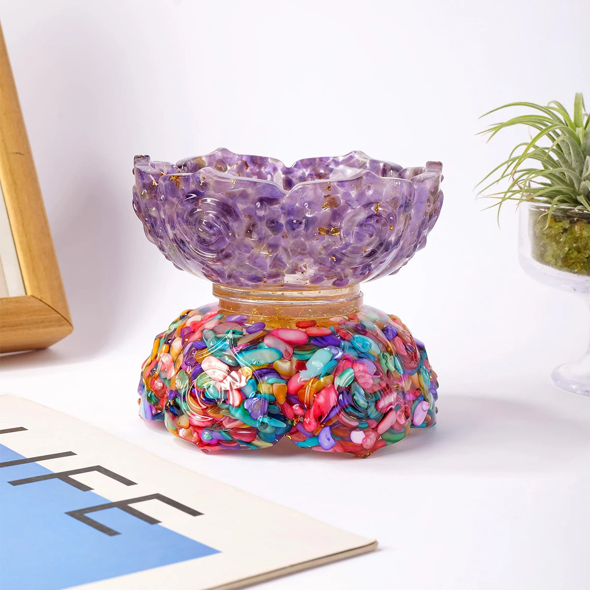 A purple crystal lotus bowl sits atop a glass base, surrounded by colorful floral crystals on a white surface.
